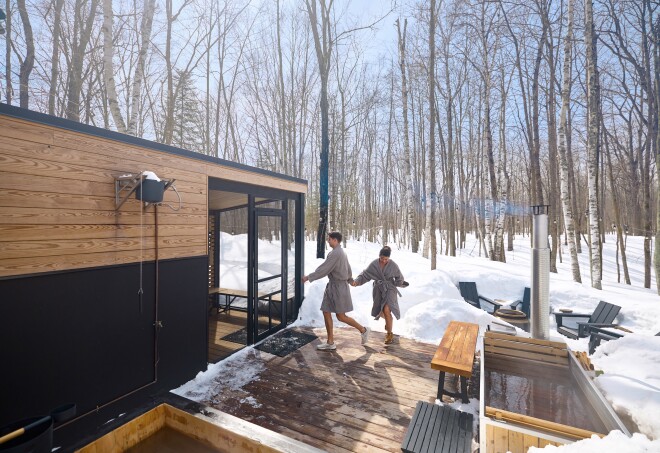 Two people wearing robes on a deck outside of a sauna surrounded by trees and snow in Minnesota.