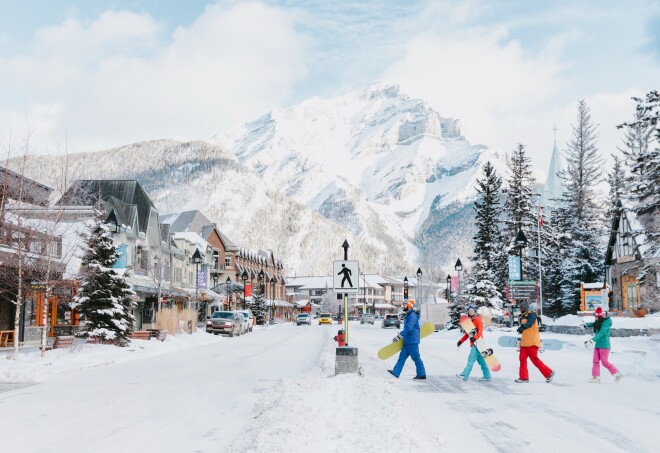 A group of people in Alberta, Banff dressed in colorful snowboarding gear town walking across a snowy street in front of Cascade Mountain.