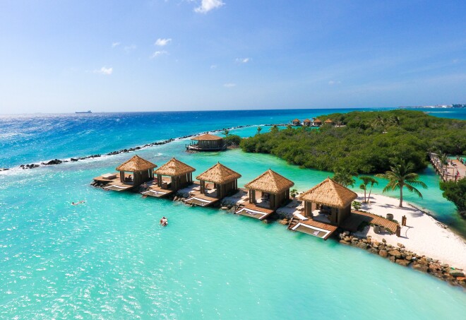 A panoramic, high view of shade structures with thatched palm roofs on a thin peninsula of white sand surrounded by the ocean at Renaissance Island, Aruba.