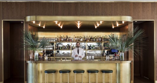 A bartender in a white suit and black tie behind a stylish golden bar with five stools and palm fronds on either side 