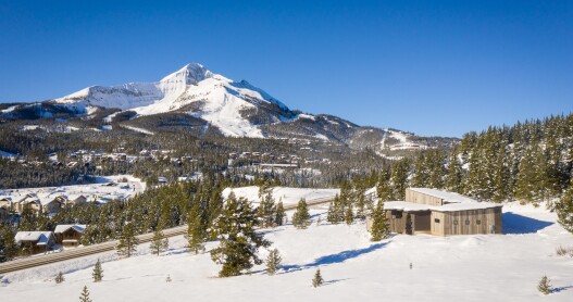 A wooden chalet surrounded by snow and evergreen trees with a snow-covered peak in the distance and bright blue skies