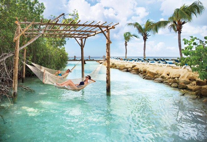 People lounging in  hammocks suspended by a wooden structure over the ocean, just off the shore in Renaissance Wind Creek Resort, Aruba