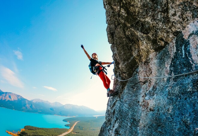 A person anchored with climbing gear to a cliff face, leaning back over the Fox Via Ferrata mountain in Alberta.