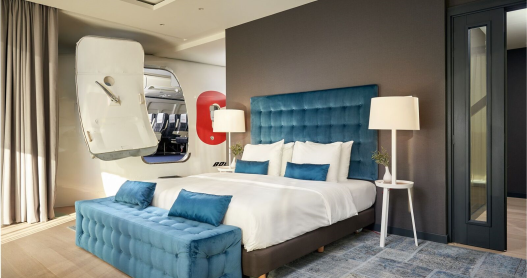 A bed with a teal blue plush headboard and foot bench in a hotel room. to the side is part of a 737 airplane that is fitted inside the suite of Corendon Amsterdam New-West hotel 